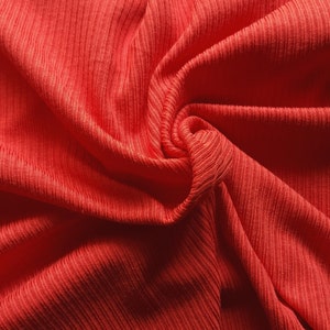 May include: Close-up of a vibrant coral-colored fabric with a ribbed texture. The material is draped and swirled, creating soft folds and shadows. The fabric's texture is emphasized by the play of light and shadow, highlighting the vertical ribbing.