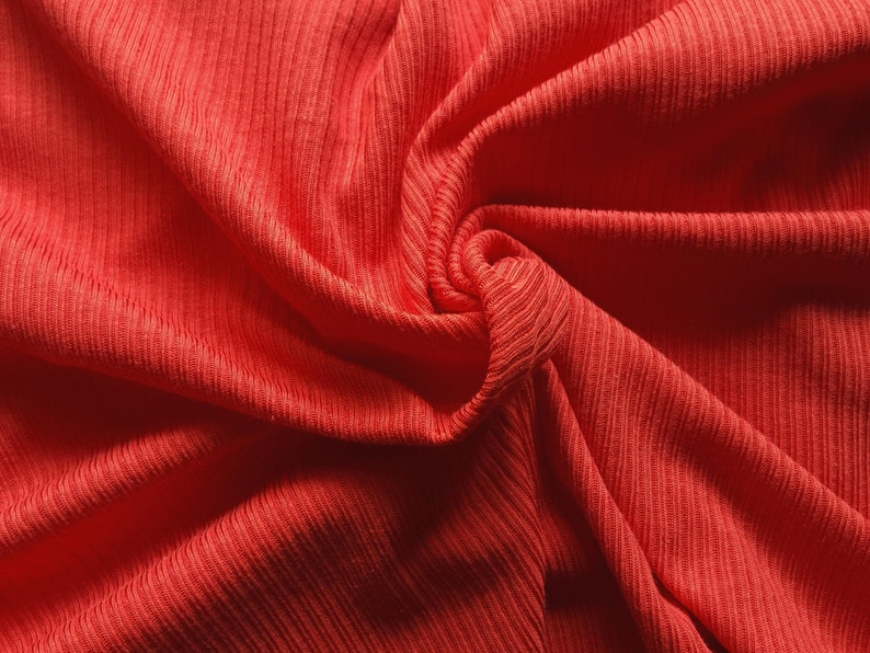May include: Close-up of a vibrant coral-colored fabric with a ribbed texture. The material is draped and swirled, creating soft folds and shadows. The fabric's texture is emphasized by the play of light and shadow, highlighting the vertical ribbing.