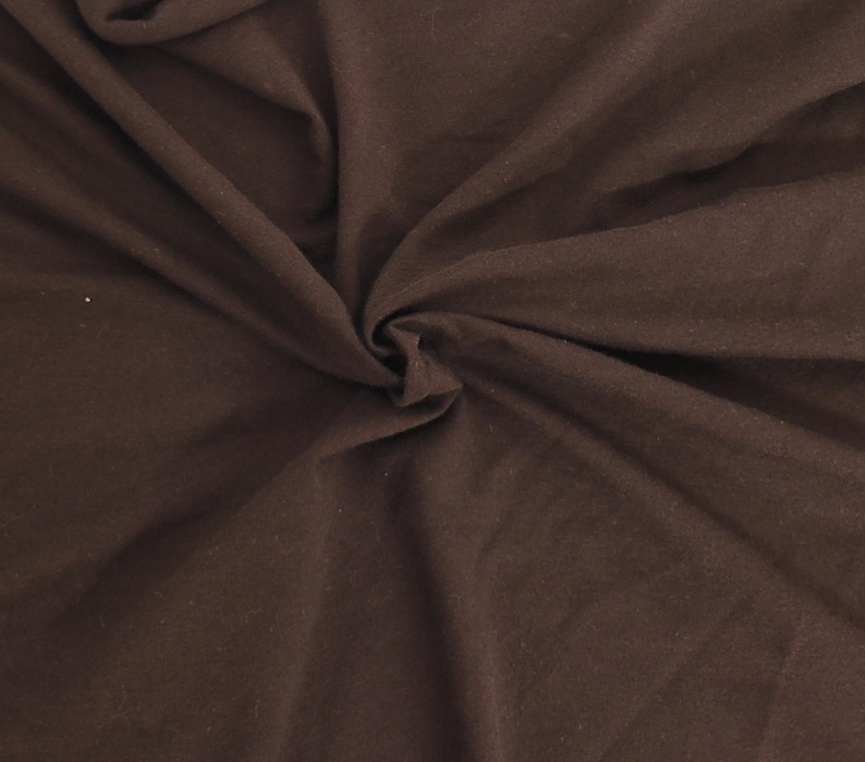 May include: A solid dark brown fabric with a soft, matte texture. The material is gathered in the center, creating natural folds and drapes that radiate outward, showcasing the consistent deep brown color and the smooth, opaque surface of the textile.