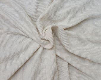 May include: Close-up of a cream-colored waffle-weave fabric. The fabric is folded and draped, creating soft shadows and texture. The weave pattern is visible, adding visual interest to the textile. The overall look is neutral and inviting.