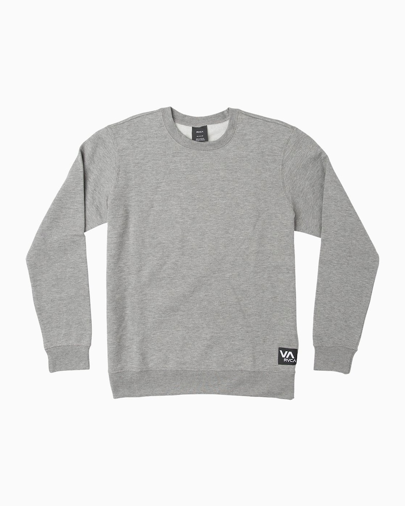 May include: A heather gray crew neck sweatshirt with long sleeves. The sweatshirt features a small black and white logo patch on the lower left side and a tag on the back of the neck. The fabric appears to be a soft, knit material.