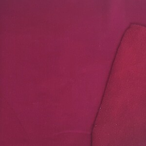 May include: A close-up of a piece of fabric in a deep burgundy color. The fabric has a smooth, matte finish. A smaller, textured piece of the same color fabric is placed diagonally across the corner of the larger piece.