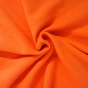 May include: A vibrant, solid orange fabric with a soft, matte texture. The material is gathered in the center, creating deep, flowing folds and natural shadows that highlight the consistent, bright orange color across the entire surface.