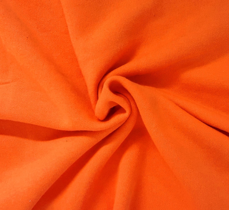 May include: A vibrant, solid orange fabric with a soft, matte texture. The material is gathered in the center, creating deep, flowing folds and natural shadows that highlight the consistent, bright orange color across the entire surface.