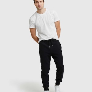 May include: A person wearing a white crew neck t-shirt, black joggers with a drawstring, and white sneakers. The outfit is casual and comfortable, suitable for everyday wear. The background is a solid, light gray.