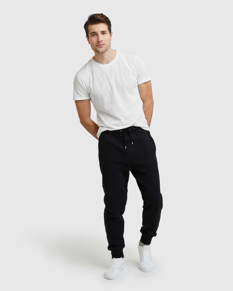 May include: A person wearing a white crew neck t-shirt, black joggers with a drawstring, and white sneakers. The outfit is casual and comfortable, suitable for everyday wear. The background is a solid, light gray.