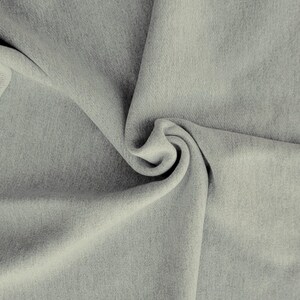 May include: Close-up of a light gray fabric with a soft, textured appearance. The fabric is draped and folded, creating a swirling pattern. The material appears to be a medium-weight textile, possibly suitable for apparel or home decor.