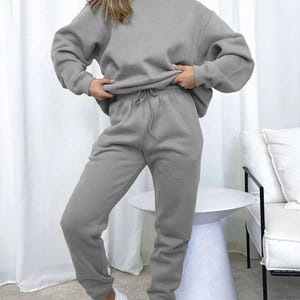 May include: A light gray sweatshirt and sweatpants set. The sweatshirt has a crew neck and long sleeves. The sweatpants have a drawstring waist and cuffed ankles. The model is wearing white sneakers.