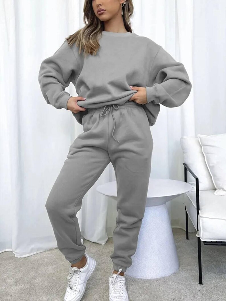 May include: A light gray sweatshirt and sweatpants set. The sweatshirt has a crew neck and long sleeves. The sweatpants have a drawstring waist and cuffed ankles. The model is wearing white sneakers.