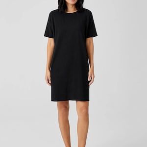 Puede incluir: Un vestido tipo camiseta de color negro s&oacute;lido que llega hasta la rodilla, con un cuello redondo sencillo y mangas cortas. La prenda presenta una silueta recta y relajada que cae sobre las rodillas, confeccionada en tela lisa y opaca.