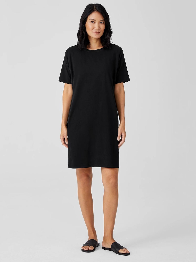 Puede incluir: Un vestido tipo camiseta de color negro s&oacute;lido que llega hasta la rodilla, con un cuello redondo sencillo y mangas cortas. La prenda presenta una silueta recta y relajada que cae sobre las rodillas, confeccionada en tela lisa y opaca.