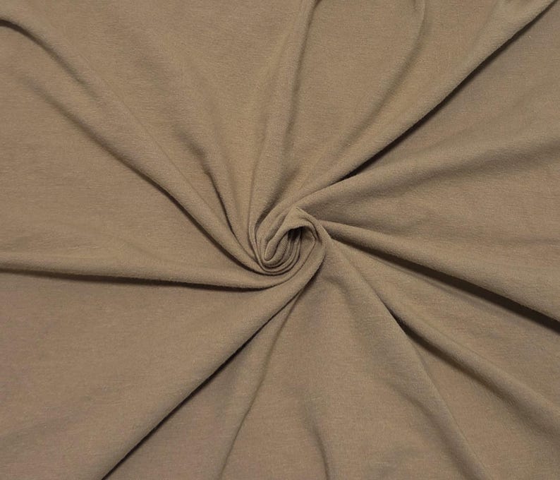 May include: A solid light brown fabric with a soft, matte texture, displayed with a central swirl that creates natural folds and drapes across the surface, highlighting the consistent, neutral earth-toned color throughout the material.