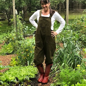 Pu&ograve; includere: Una donna indossa una salopette verde oliva con una camicia bianca a maniche lunghe e stivali da pioggia rossi in piedi in un giardino.