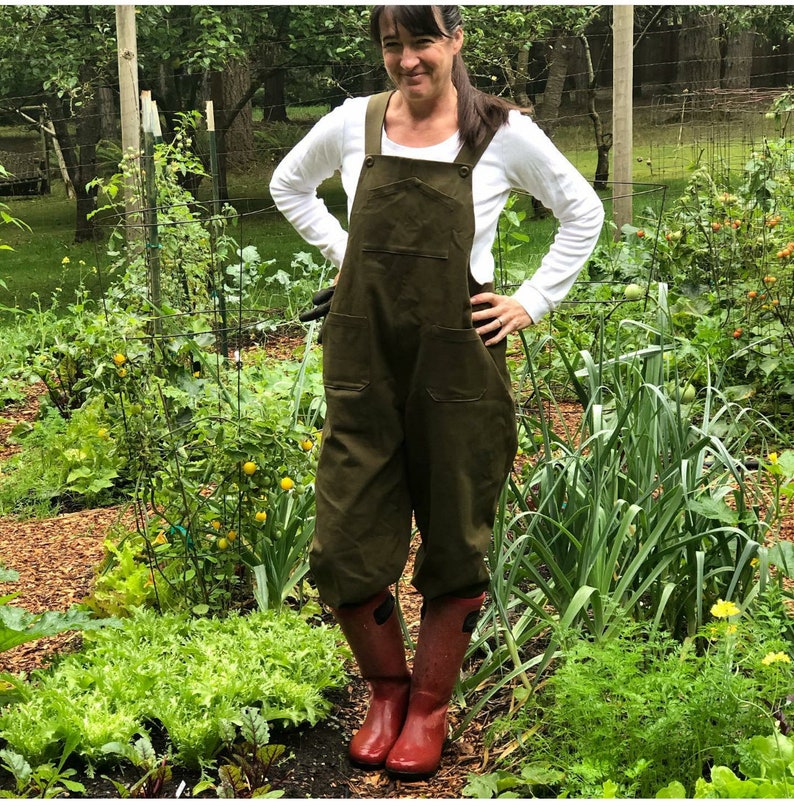 Pu&ograve; includere: Una donna indossa una salopette verde oliva con una camicia bianca a maniche lunghe e stivali da pioggia rossi in piedi in un giardino.