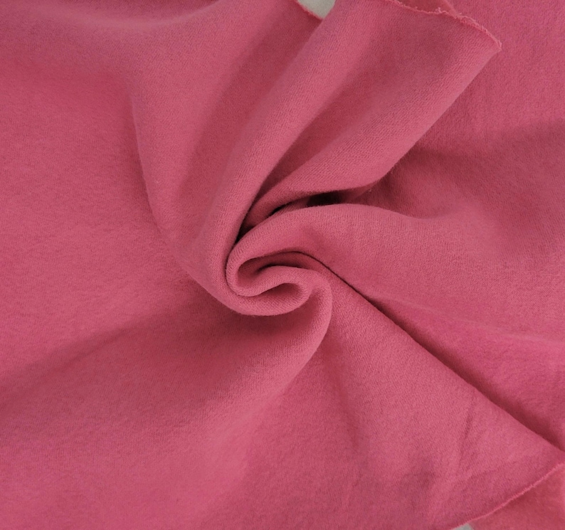 May include: A solid, deep rose-pink fabric with a soft, matte, and slightly fuzzy texture. The material is draped in a central swirl, creating natural folds and shadows that highlight the consistent, uniform color and smooth surface of the textile.