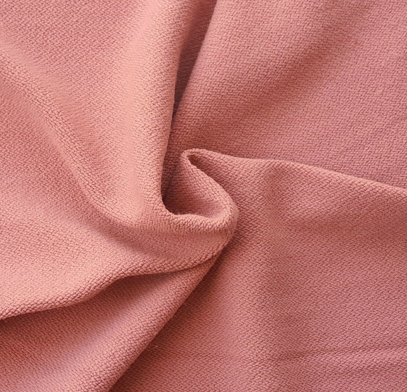 May include: Close-up of a soft, pink fabric with a textured surface. The fabric is folded and draped, showing its texture and drape.