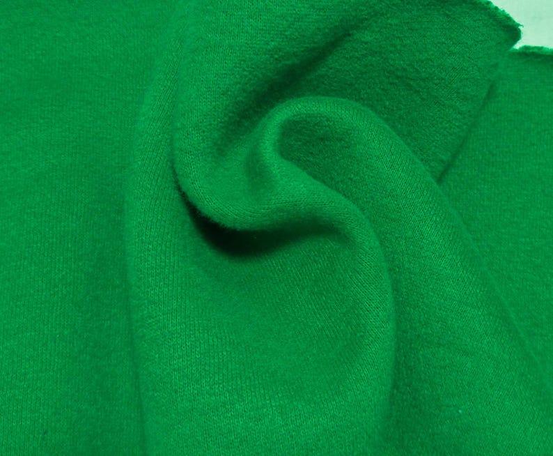 May include: A vibrant, solid green fabric with a soft, slightly fuzzy texture. The material is draped to create gentle folds and curves, highlighting the consistent, saturated emerald green color across the entire surface of the textile.