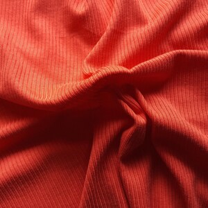 May include: Close-up of a vibrant orange fabric with a ribbed texture. The material is draped and folded, creating shadows and highlights that emphasize the texture and color. The fabric appears soft and possibly knitted.