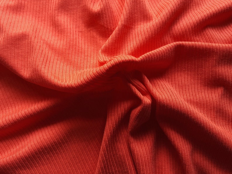 May include: Close-up of a vibrant orange fabric with a ribbed texture. The material is draped and folded, creating shadows and highlights that emphasize the texture and color. The fabric appears soft and possibly knitted.