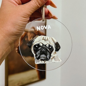 May include: A round, clear acrylic ornament with a hand-painted portrait of a pug dog. The ornament has a white string for hanging and the name "NOVA" written in white paint at the top.