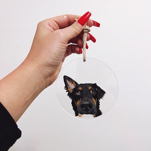 May include: A round, clear acrylic bauble with a painted portrait of a black and brown dog with a white patch on its chest. The dog is looking at the camera with its head tilted to the side. The bauble has a beige string attached to it.