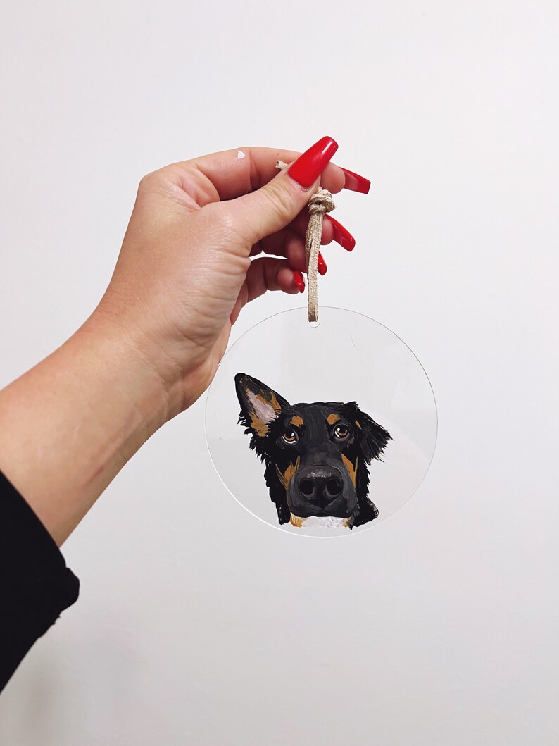 May include: A round, clear acrylic bauble with a painted portrait of a black and brown dog with a white patch on its chest. The dog is looking at the camera with its head tilted to the side. The bauble has a beige string attached to it.