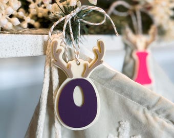 May include: A purple felt reindeer ornament with a white outline and antlers. The ornament is hanging from a white stocking with a snowflake design.