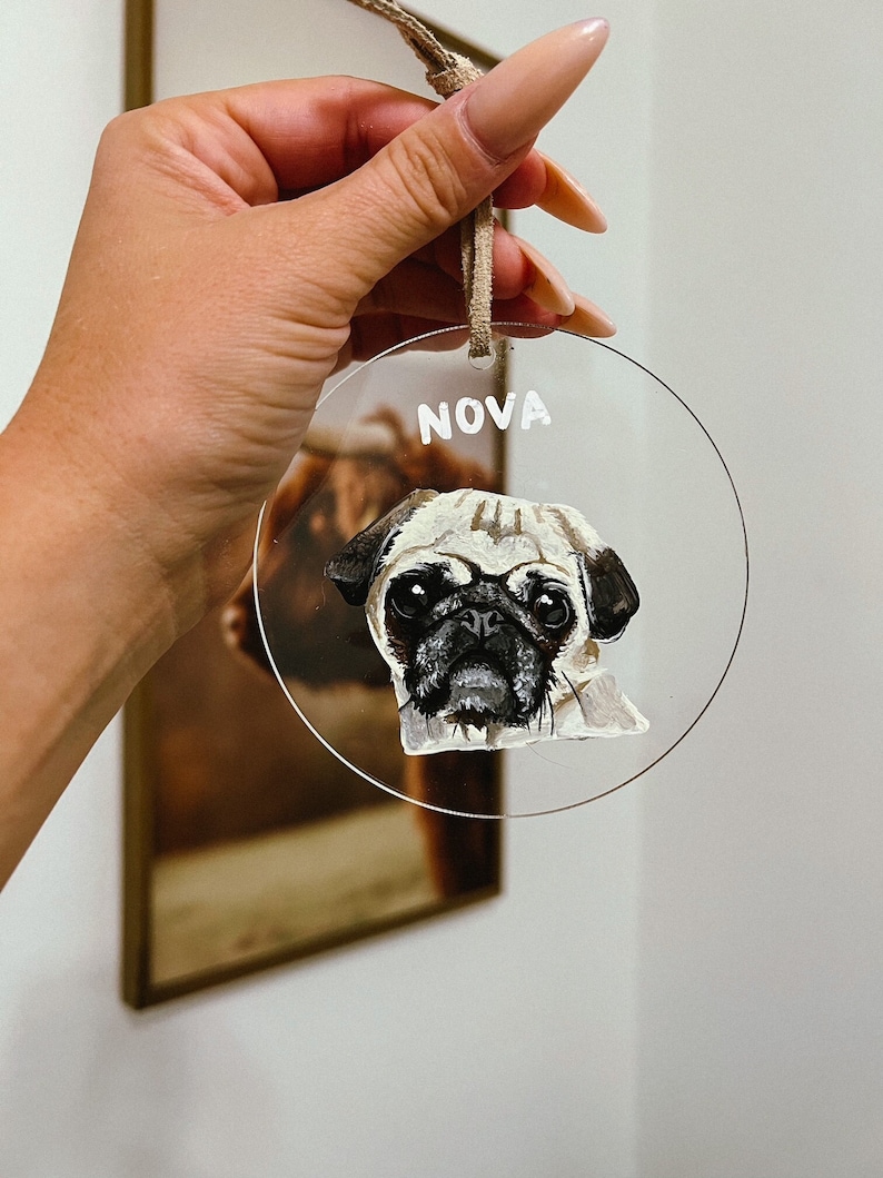 May include: A round, clear acrylic bauble with a hand-painted portrait of a pug dog. The bauble has a white string for hanging and the name "NOVA" written in white paint at the top.