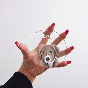 May include: A round, clear acrylic bauble with a painted portrait of a brown and white dog. The dog's name, "ARTIE", is written in white on the bauble.