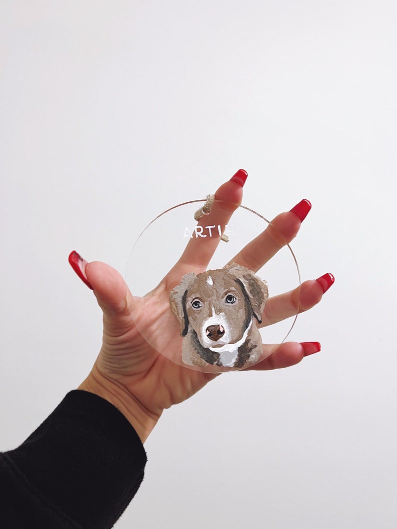 May include: A round, clear acrylic bauble with a painted portrait of a brown and white dog. The dog's name, "ARTIE", is written in white on the bauble.