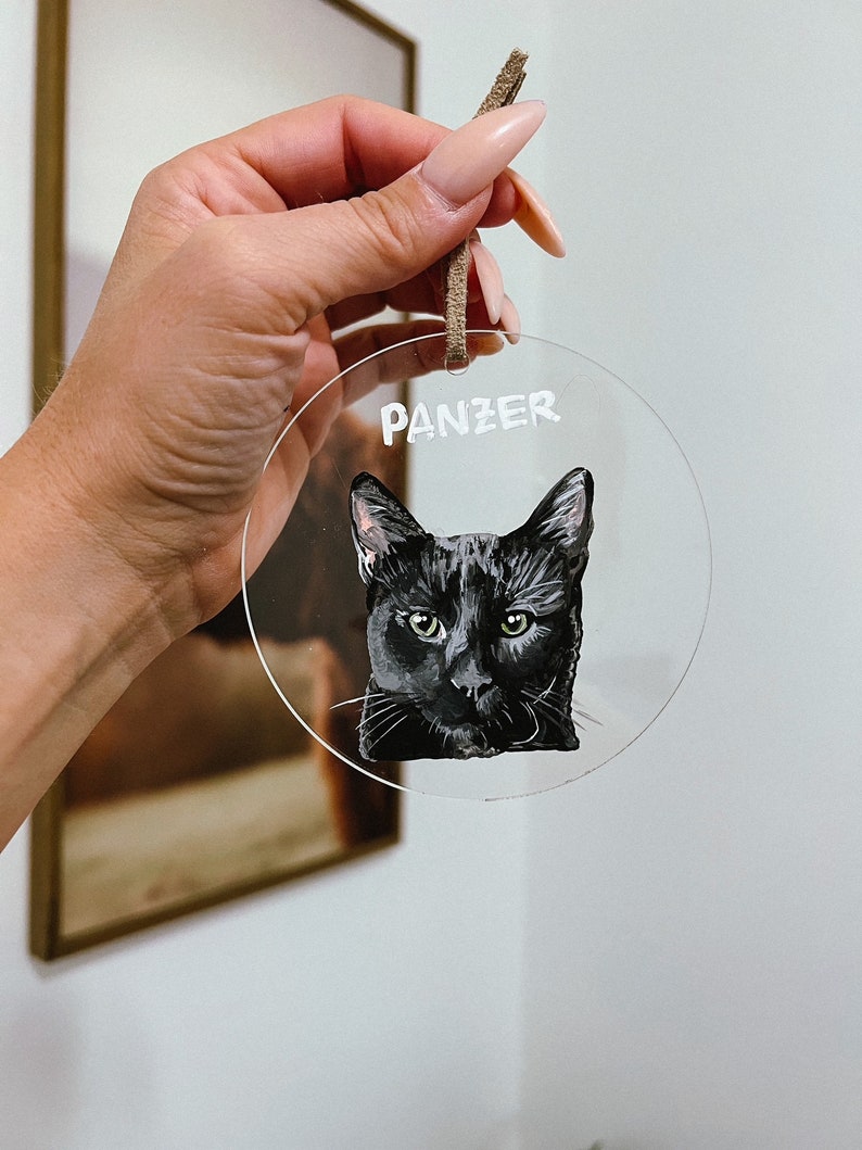 May include: A clear round bauble with a black cat painted on it. The cat's name, "Panzer", is written in white lettering at the top of the bauble.