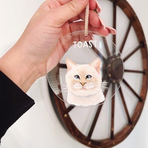 May include: A clear round bauble with a painted portrait of a white cat with blue eyes. The bauble has a string for hanging and the word "TOAST" written on it.