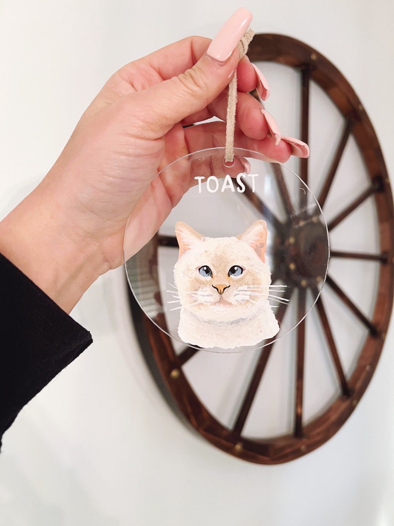 May include: A clear round bauble with a painted portrait of a white cat with blue eyes. The bauble has a string for hanging and the word "TOAST" written on it.