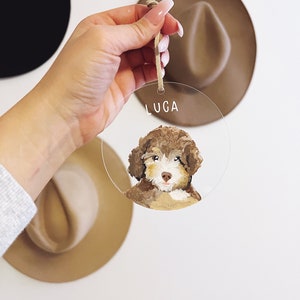 May include: A round, clear acrylic bauble with a painted portrait of a brown and white dog. The bauble has a hole at the top for hanging and the name "LUGA" is written in white lettering at the top.