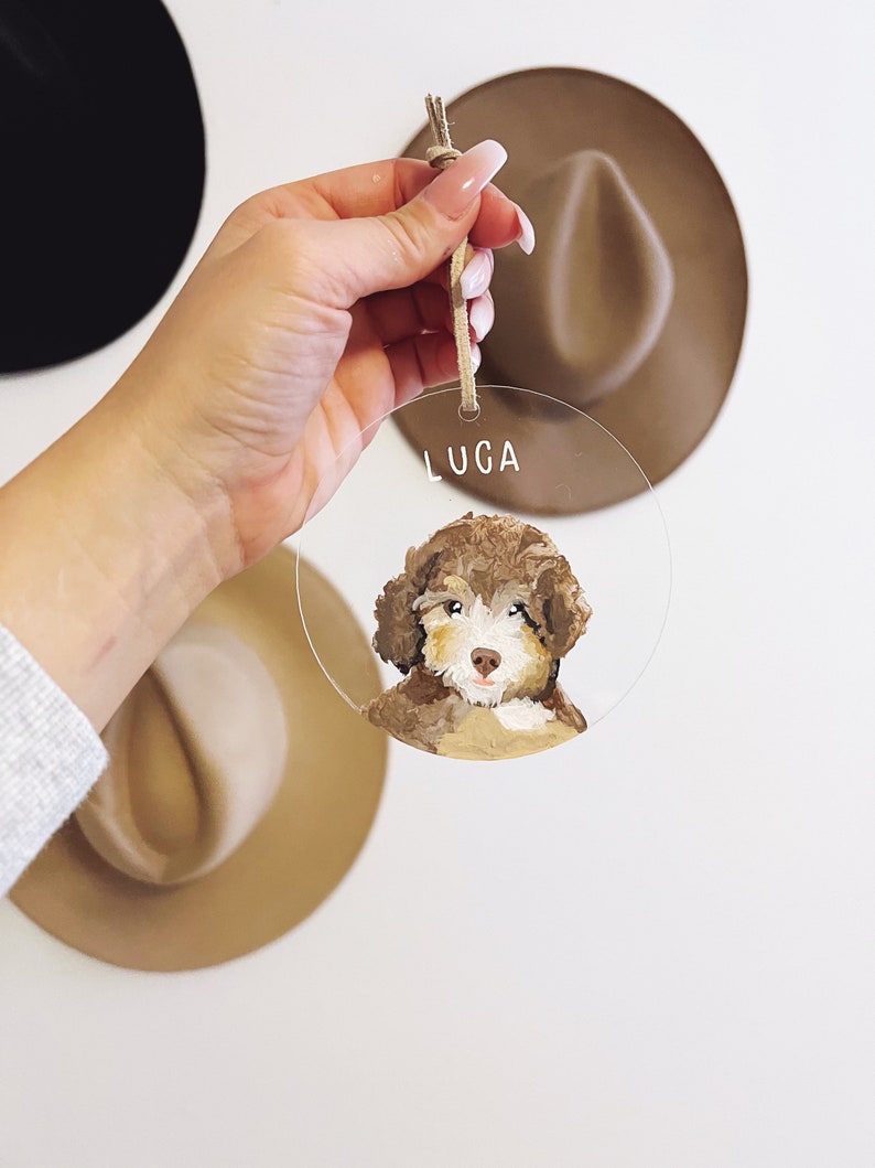 May include: A round, clear acrylic bauble with a painted portrait of a brown and white dog. The bauble has a hole at the top for hanging and the name "LUGA" is written in white lettering at the top.