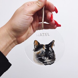 May include: A round, clear acrylic bauble with a painted portrait of a calico cat. The bauble has a brown string for hanging and the name "Hazel" written on the front.
