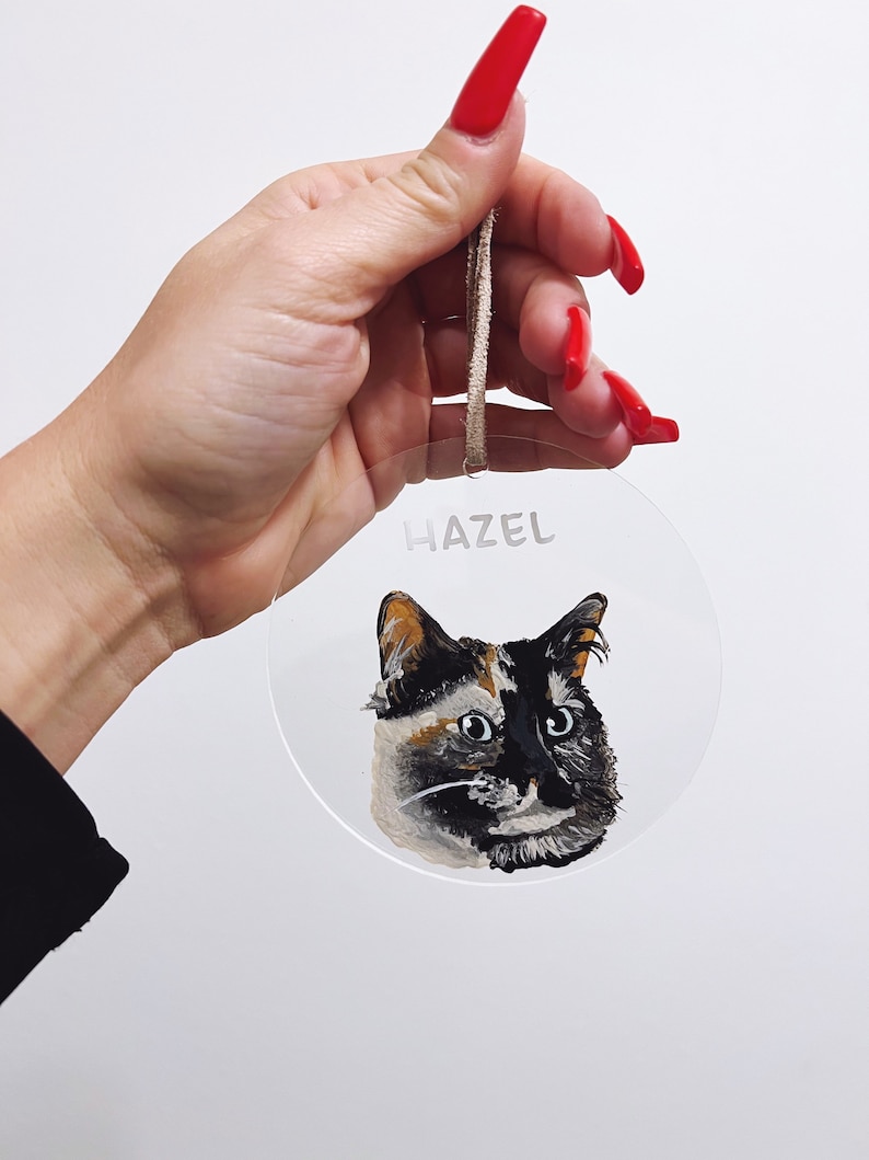 May include: A round, clear acrylic bauble with a painted portrait of a calico cat. The bauble has a brown string for hanging and the name "Hazel" written on the front.