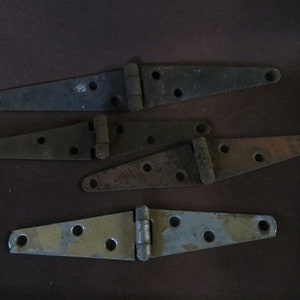 May include: Four vintage metal hinges in various shades of gray and brown. Each hinge has a rectangular shape with rounded ends and multiple screw holes. The hinges are arranged on a dark brown surface.