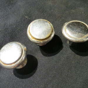 Può includere: Tre pomelli per mobili argentati dal design circolare. I pomelli hanno una parte superiore leggermente a cupola e una base corta e cilindrica. Il metallo mostra segni di usura e un po' di ruggine sui bordi, suggerendo che siano vintage o antichi.