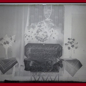 May include: An antique photograph featuring a casket, flanked by floral arrangements in vases and on small tables. A large floral arrangement hangs on the wall behind the casket. The image is in black and white.