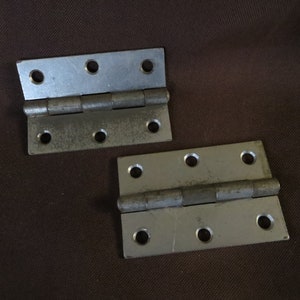 May include: Two metal hinges with a silver finish, each with four screw holes. The hinges are slightly weathered, showing signs of use. They are set against a brown background.