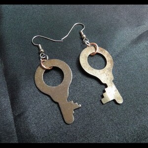 May include: A pair of handmade earrings featuring a key design. The earrings are made of silver-colored metal with a copper-colored ring connecting the key to the ear wire. The keys have a vintage, rustic appearance.