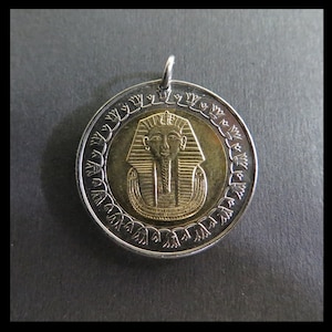May include: A circular pendant featuring a gold-toned center embossed with the bust of an Egyptian pharaoh, surrounded by a silver-toned border decorated with a repeating lotus flower pattern and a small metal loop at the top for attachment.