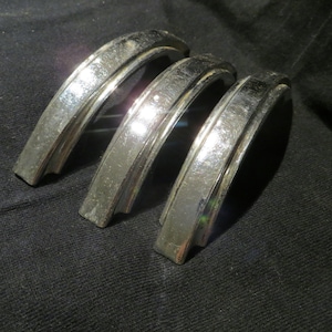 Op de afbeelding: Drie vintage chromen kastgrepen met een gebogen, gewelfd ontwerp. De grepen hebben een glanzend, reflecterend oppervlak en zijn op een donkere achtergrond geplaatst. Deze hardwarestukken zijn waarschijnlijk voor een dressoir of kast.