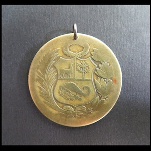 Könnte beinhalten: Ein runder, antik goldfarbener Anhänger mit einem detaillierten geprägten Design. Der Anhänger zeigt ein zentrales Schild mit einem Hirsch, einem Baum und einem Füllhorn, umgeben von Lorbeerzweigen und floralen Elementen. Oben befindet sich eine kleine Schlaufe zum Aufhängen.