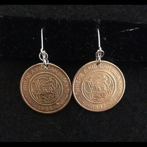 May include: A pair of coin-shaped earrings. Each earring features a cartoon character wearing a hat, with the text "WHERE A KID CAN BE A KID" and the years 1996 and 1997. The earrings have silver-colored hooks.