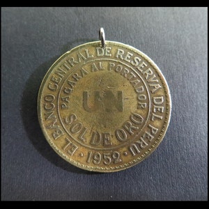 Könnte beinhalten: Ein goldfarbener Münzanhänger mit einer Schlaufe oben. Die Münze trägt spanischen Text, darunter "BANCO CENTRAL DE RESERVA DEL PERU" und "UN SOL DE ORO 1952". Die Münze hat ein abgenutztes Aussehen.
