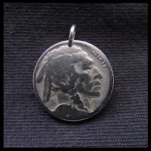 May include: A silver pendant made from a coin, depicting a detailed profile of a Native American chief. The word "LIBERTY" is visible. The pendant has a small loop for a chain.