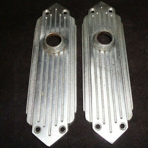 May include: Two vintage silver-colored door handle backplates. Each plate has a rectangular shape with vertical grooves and a circular opening for the handle. The top has a decorative pointed design, and the bottom has two screw holes.