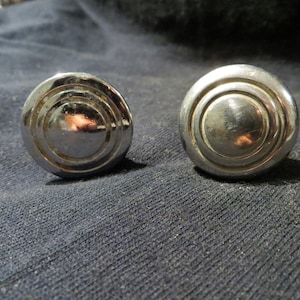 May include: Two shiny, chrome-colored cabinet knobs. Each knob has a circular design with concentric rings. The knobs are round and have a reflective surface, suggesting a polished finish. They are likely used for furniture or cabinetry.